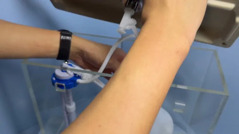 Arm reaching into a clear acrylic toilet tank to adjust a blue fill valve and white tubing