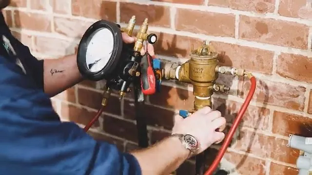Technician attaching a red-hosed test gauge to a brass backflow assembly mounted on a brick wall