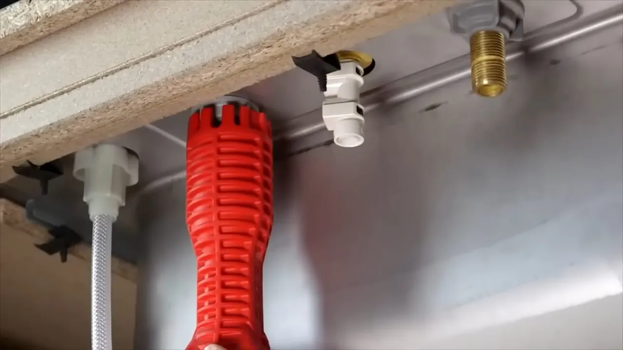 Red basin wrench reaching up under a kitchen sink counter beside shutoff valves and white supply lines