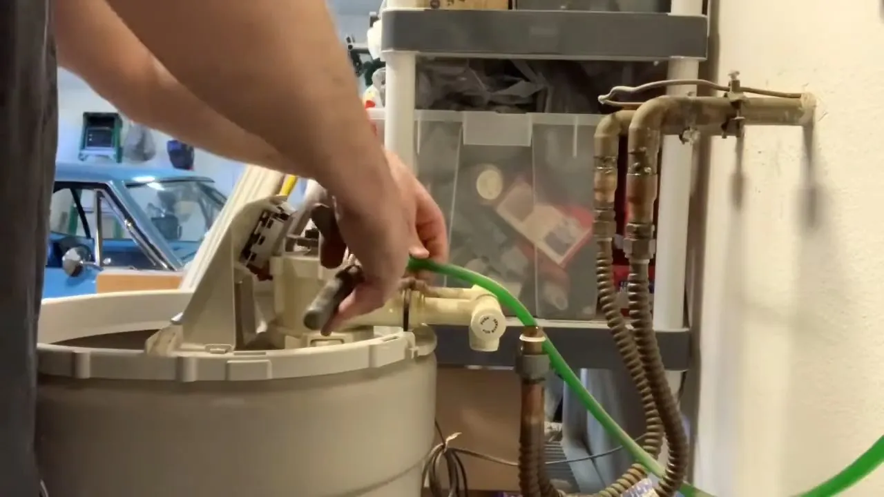 Beige softener tank with a green drain hose and brass valve lines beside a hand reaching in