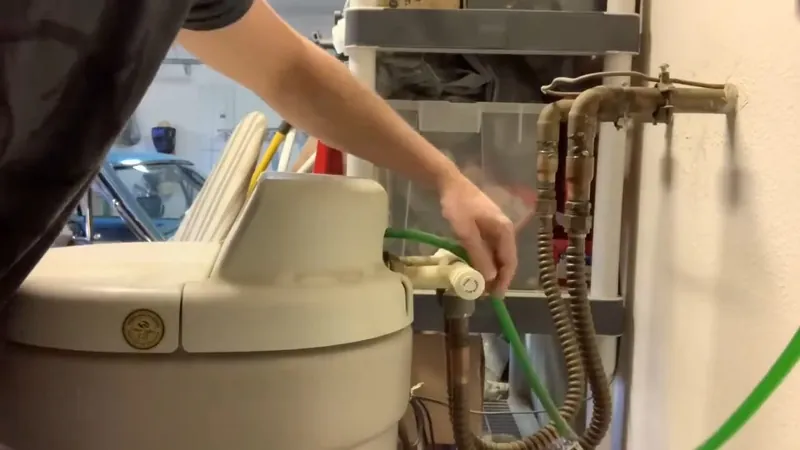 Beige water softener tank with a green hose attached and brass supply lines, with a hand at the valve