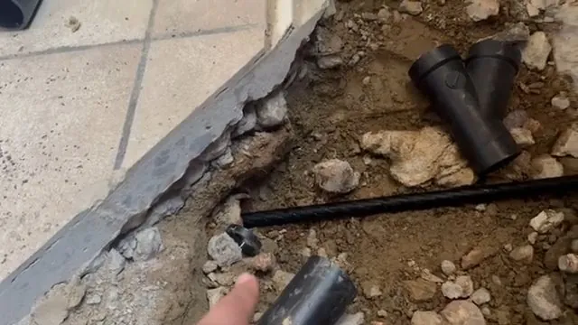 Black ABS wye fitting and pipe exposed in a broken concrete slab edge with loose rock