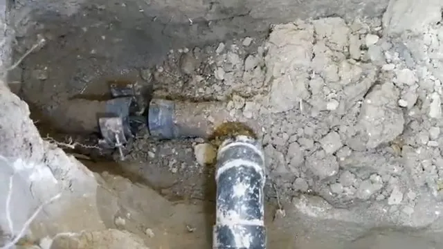 Broken drain pipe and a black fitting exposed in a trench of chipped concrete and wet soil