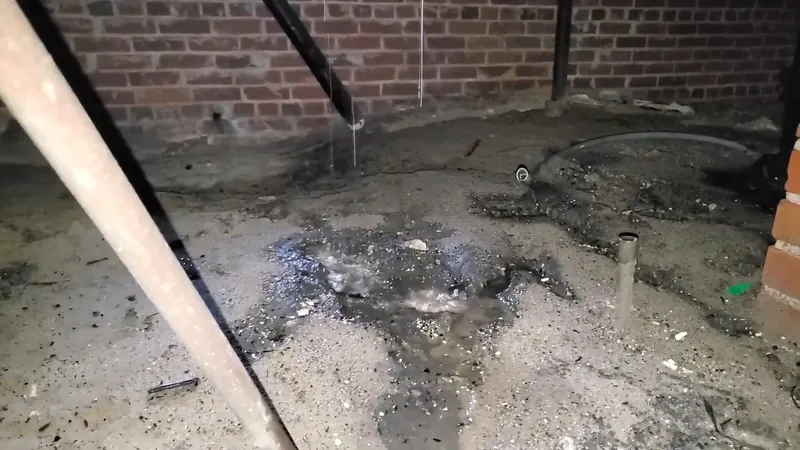 Broken concrete slab with dark wet soil and a pipe stub in front of a brick wall