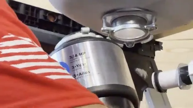Close view of a garbage disposal body and its upper flange under a sink with a drain line to the side