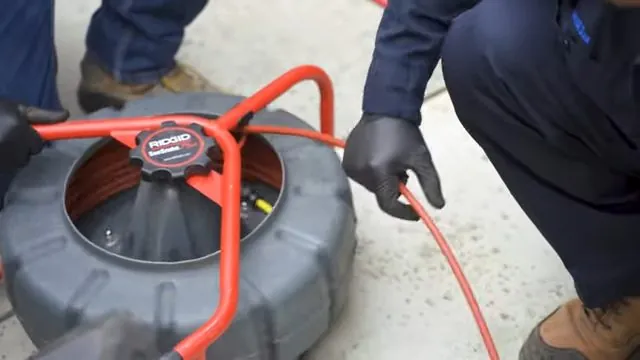 Red drum-style drain machine with cable being fed out beside a worker's boots on concrete