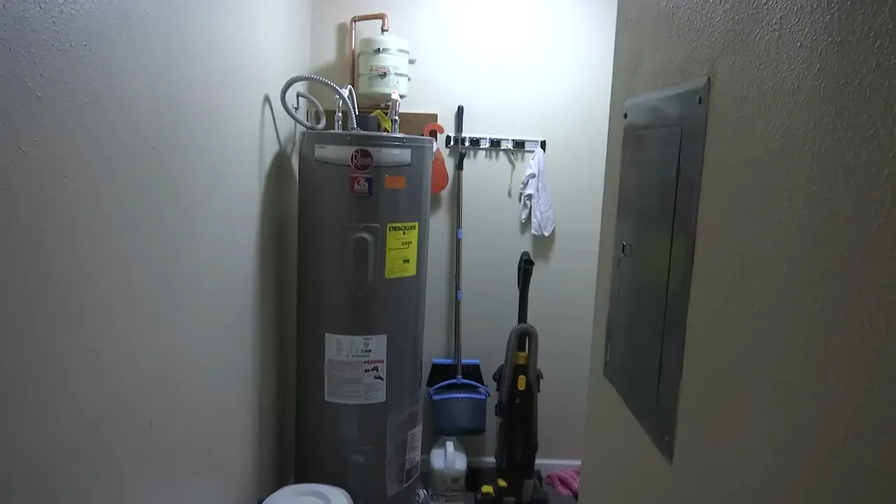 Gray electric water heater in a utility closet with a mop and tools hanging on the wall and a panel at right