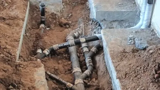 Exposed cast iron and PVC drain pipes branching in a dug trench beside a concrete foundation