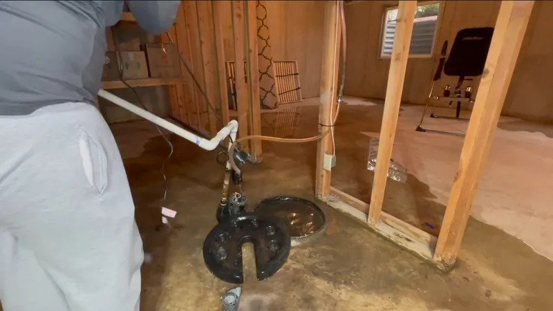 Framed basement room with a standing-water floor, a sump basin cover and a worker in the corner