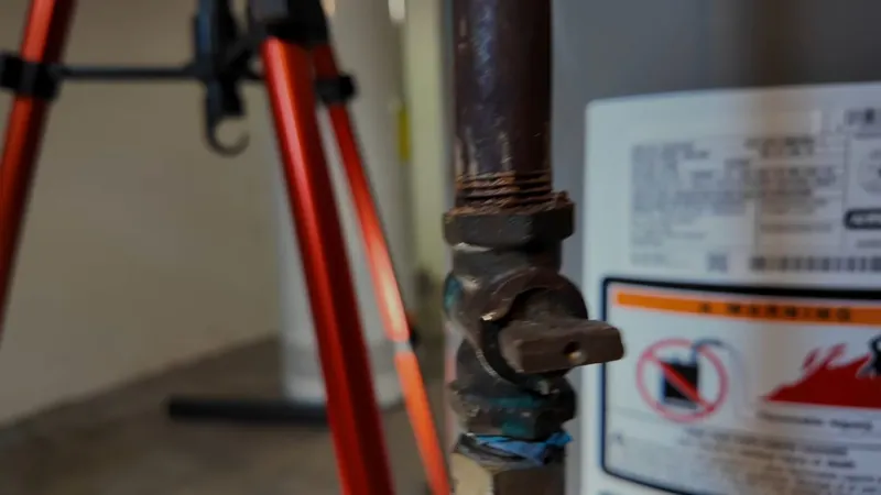 Brass gas shutoff valve on a pipe beside a water heater with a warning label and a tripod behind