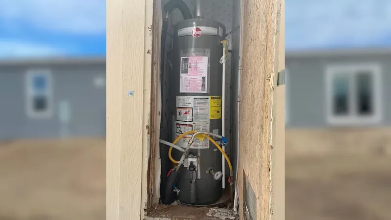 Gas water heater standing in a narrow closet with a yellow gas line, framed by blurred houses behind