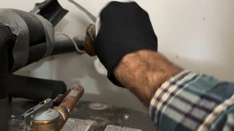 Black-gloved hand with a plaid sleeve working at copper fittings and a shutoff valve beside a tank