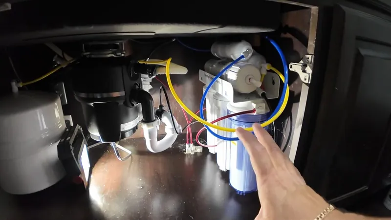Hand adjusting white and blue reverse osmosis cartridges and colored tubing in a dark sink cabinet