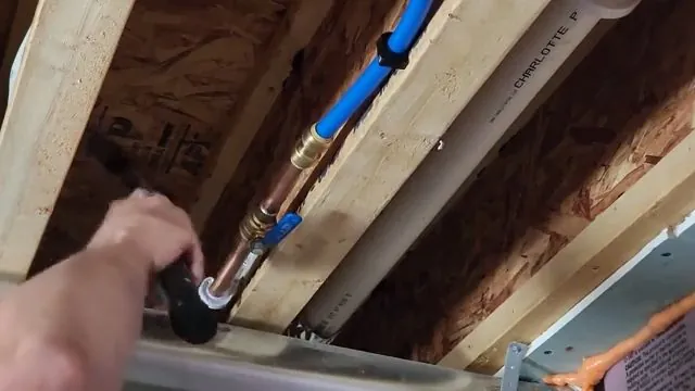 Hand pushing a copper fitting onto a blue PEX line run through framing joists with insulation above