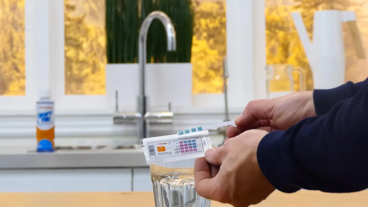 Hand dipping a water test strip into a glass beside a chrome filtered-water faucet