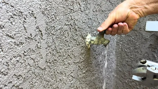 Hand opening a brass hose bib on a stucco wall with a thin stream of water running down