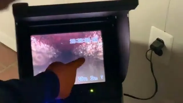 Hand pointing to a pinkish pipe interior on a sewer camera monitor with a power cord plugged into the wall