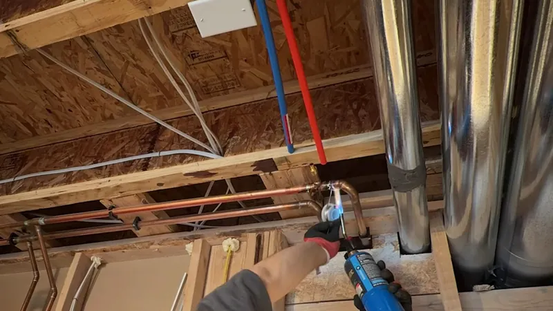 Hand with a blue tool working at red and blue PEX lines beside silver ducts under open joists