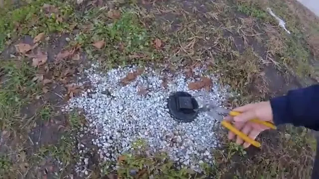 Hand with a yellow tool reaching toward a black valve cap in a patch of white gravel in a lawn