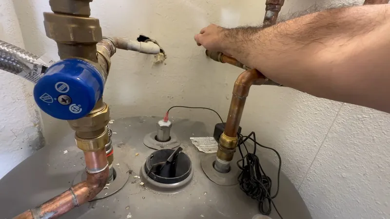 Hands connecting copper lines at the top of a gray water heater with a blue valve and an electrical cord