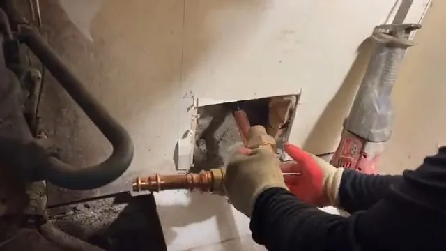 Gloved hands cutting copper supply pipe through a square opening in a wall