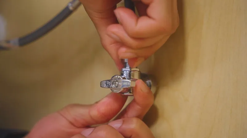 Two hands holding a chrome angle stop valve with a braided supply line against a wood panel