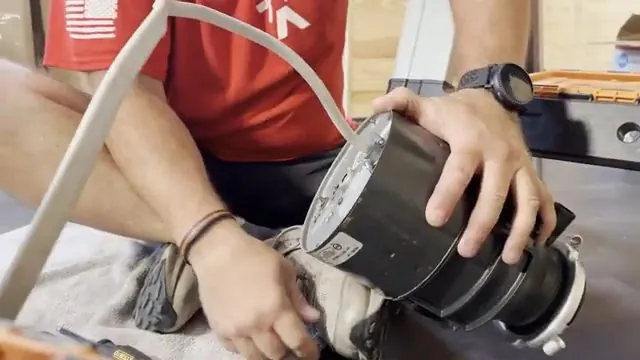 Hands lifting a black garbage disposal into position under a counter with a white cord nearby