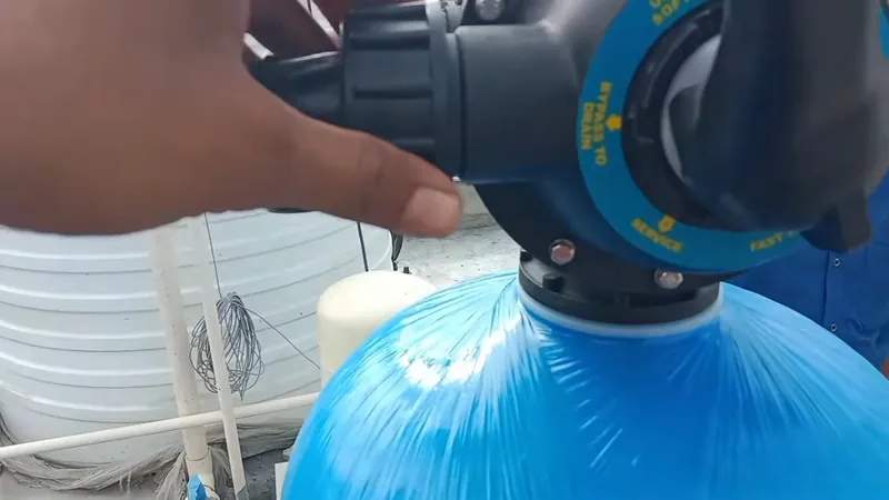 Hands on the control valve head of a blue water softener tank with a white tank behind it
