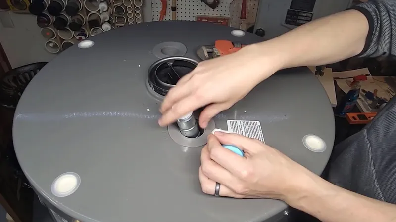 Two hands working at the fittings on top of a gray water heater tank in a workshop
