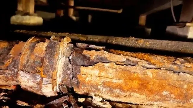 Heavily rusted pipe lying under wood floor joists with flaking orange scale along its length