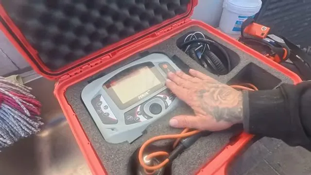 Electronic leak detection unit sitting in a red hard case with foam cutouts and a hand resting beside it