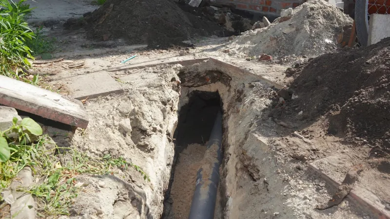 Open trench in sandy soil with black sewer pipe at the bottom and piles of dirt along the edge