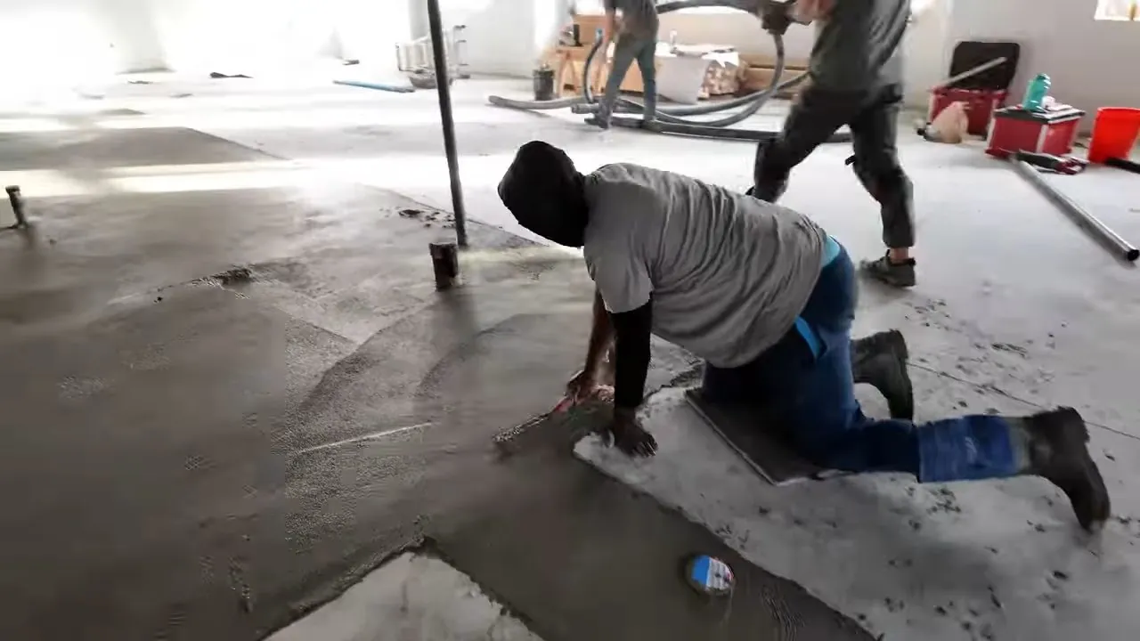 Man in a gray shirt crawling over a fresh concrete slab in an open building with workers behind him