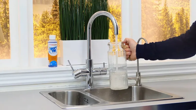 Man's hand filling a glass jar at a chrome faucet beside a double-bowl steel sink