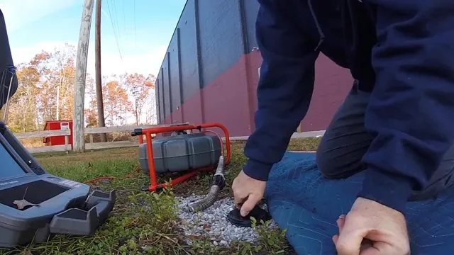 Man in a navy jacket kneeling at a lawn cleanout with a red camera drum reel beside him