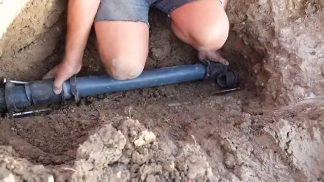 Man kneeling in a narrow dirt trench placing a blue pipe section with both hands
