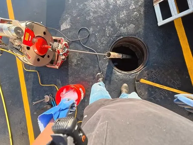 Top-down view of a worker holding a red-reeled jetter at a round manhole opening on a dark surface