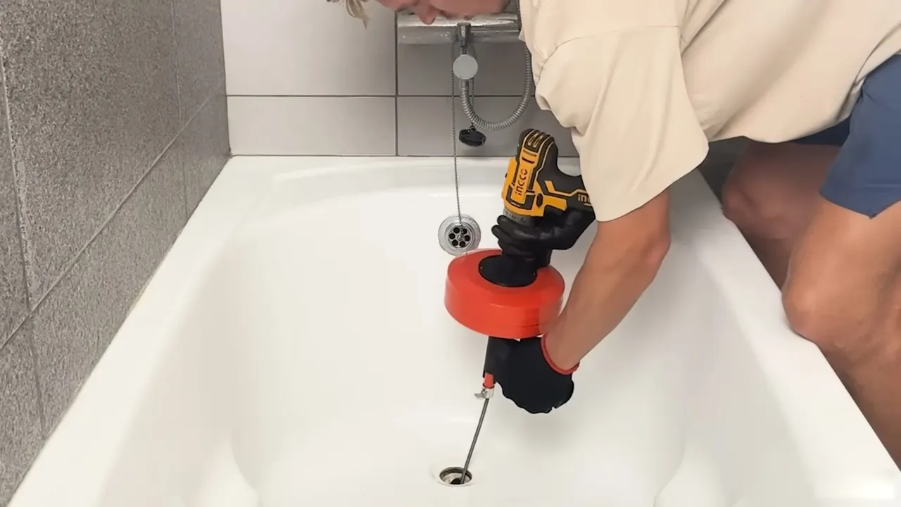Plumber using a drill-driven drain auger with an orange head fed into a white bathtub drain