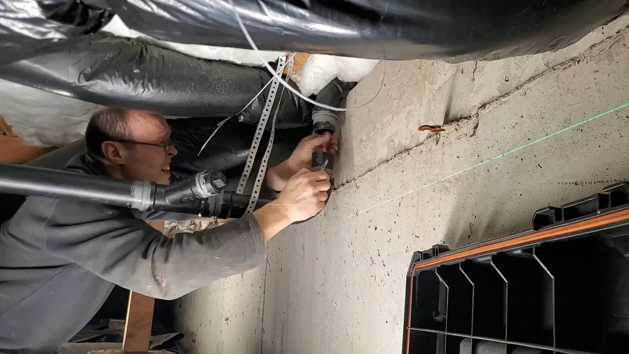 Plumber leaning across ductwork in an unfinished ceiling to work on a pipe fastened to a concrete wall
