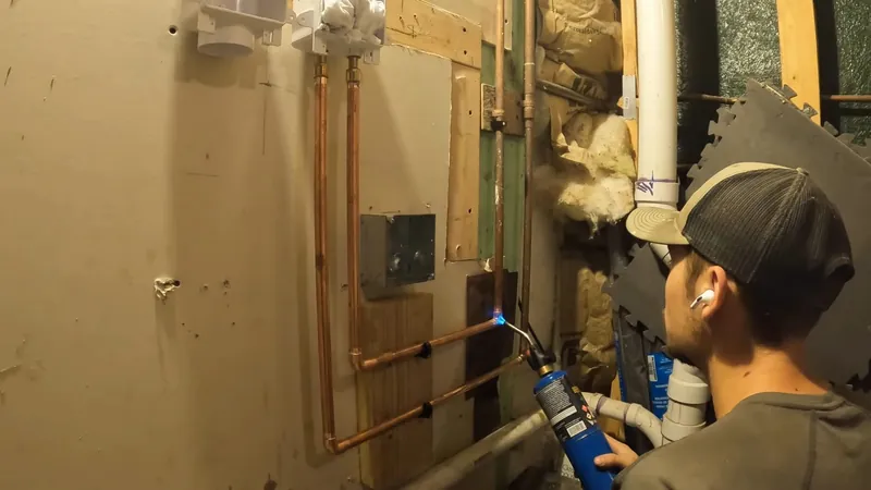 Plumber in a ballcap using a blue torch to solder copper pipes on a framed wall with insulation