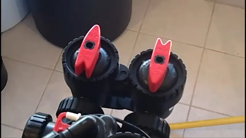 Two black valves with red handles on a softener bypass assembly over a tile floor with a yellow hose
