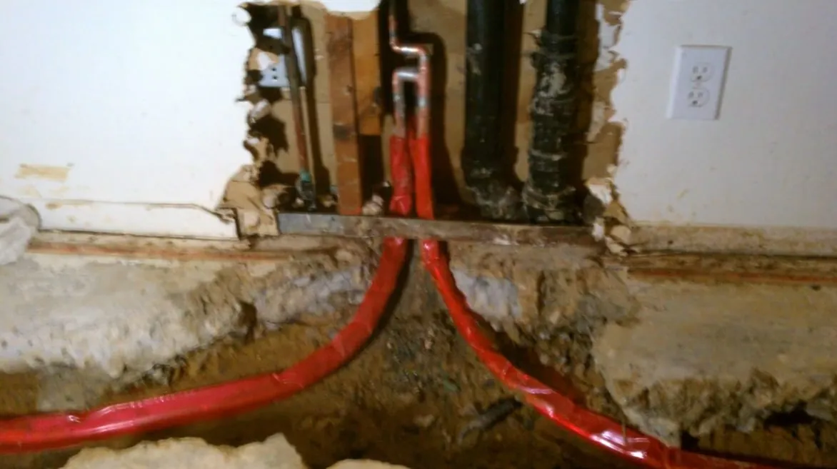Two red PEX lines running through a trench cut in a concrete slab beside a wall opening