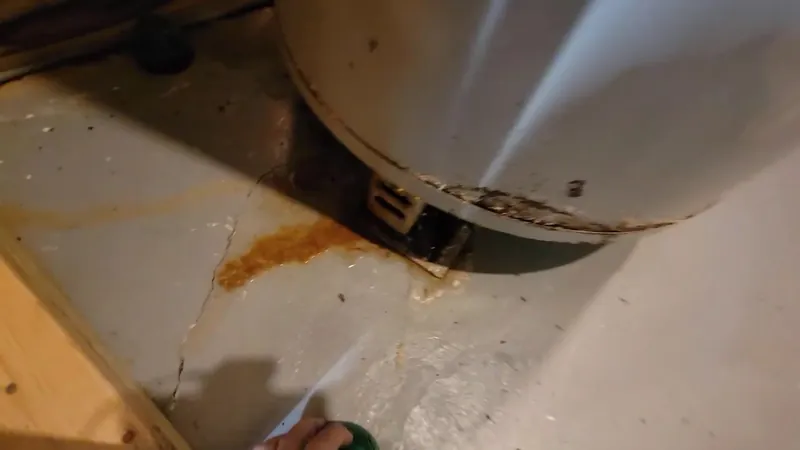 Orange rust stain and wet floor around the base of a gray water heater tank
