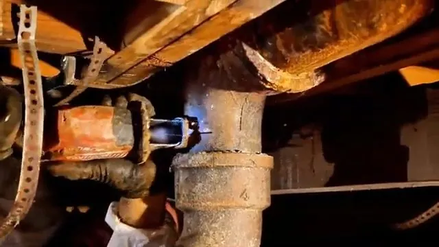 Rusty cast iron drain with an orange fitting under floor joists, lit from below by a torch