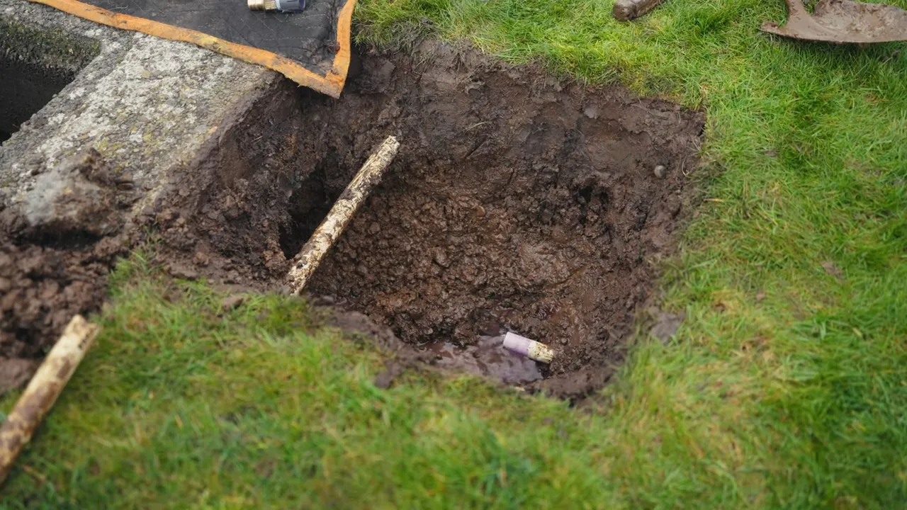 Square hole dug in a green lawn with dark soil and a stick leaning in it, next to a concrete edge