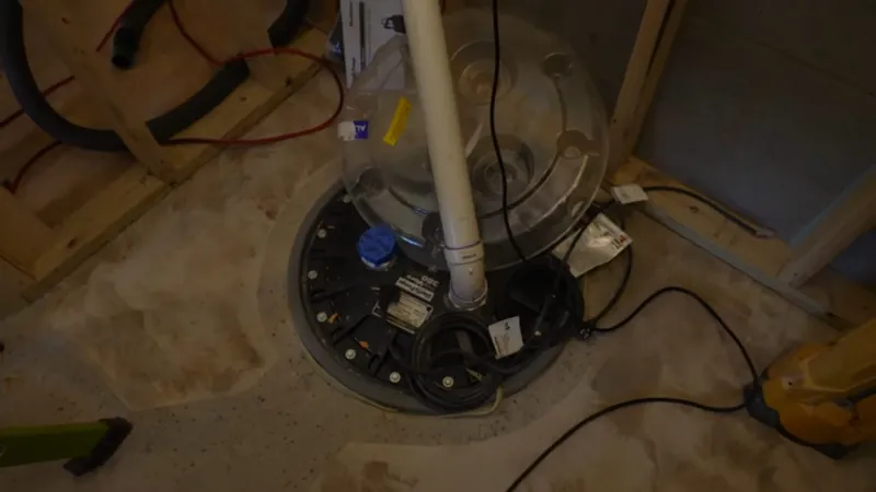Sump basin set into a basement floor with a white PVC discharge pipe and cords beside open framing