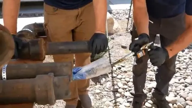 Two plumbers in work gloves cutting old cast iron drain pipe over gravel with a snap cutter
