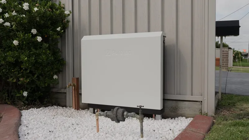 White metal cabinet mounted on an exterior wall above a bed of white gravel with pipes below