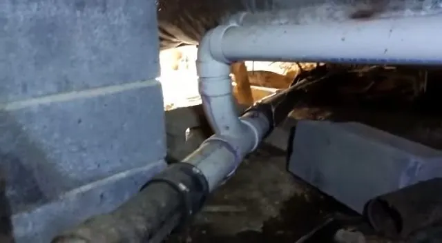 White PVC and black drain pipes running along a block wall in a crawl space over wet ground