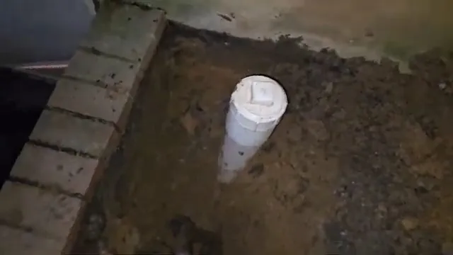White PVC cleanout riser standing in dug dirt beside a brick edge